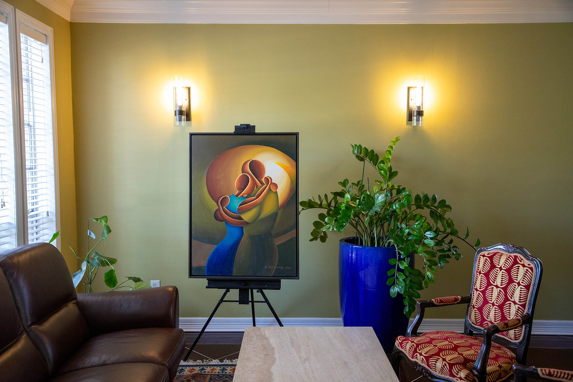 A cozy green-walled room includes the Love Me Tender - Canvas Art on an easel, showing an abstract embrace in contemporary African style. It features a brown leather sofa, a red-patterned chair, a marble table, and a large blue pot with a green plant. Two wall sconces light up the area.