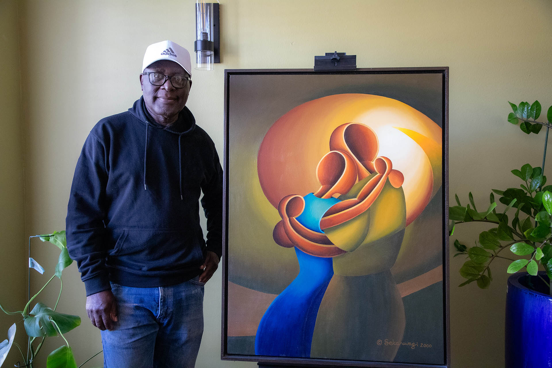 A person in a black hoodie and white cap stands by Love Me Tender - Canvas Art, featuring warm-toned abstract figures, perfectly complementing the surrounding greenery and exuding inspirational interior design vibes.