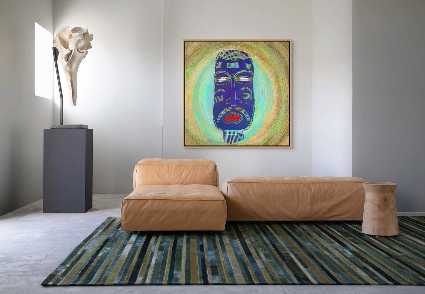 A modern living room showcases a tan leather sofa and striped rug, with a nearby wooden stool. The wall is adorned by the Blue Mask canvas art, created by an African artist, and a sculpture resembling a large bone stands on a pedestal.