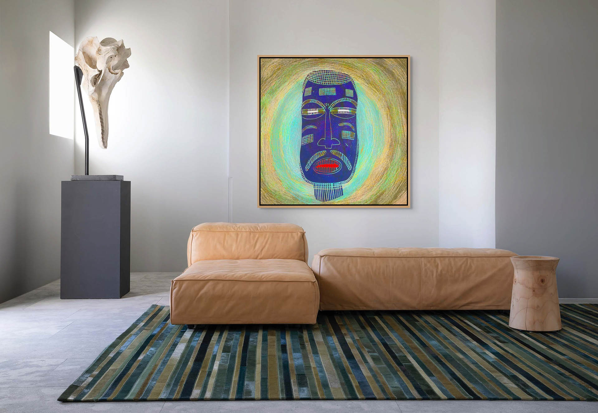 A modern living room showcases a tan leather sofa and striped rug, with a nearby wooden stool. The wall is adorned by the Blue Mask canvas art, created by an African artist, and a sculpture resembling a large bone stands on a pedestal.