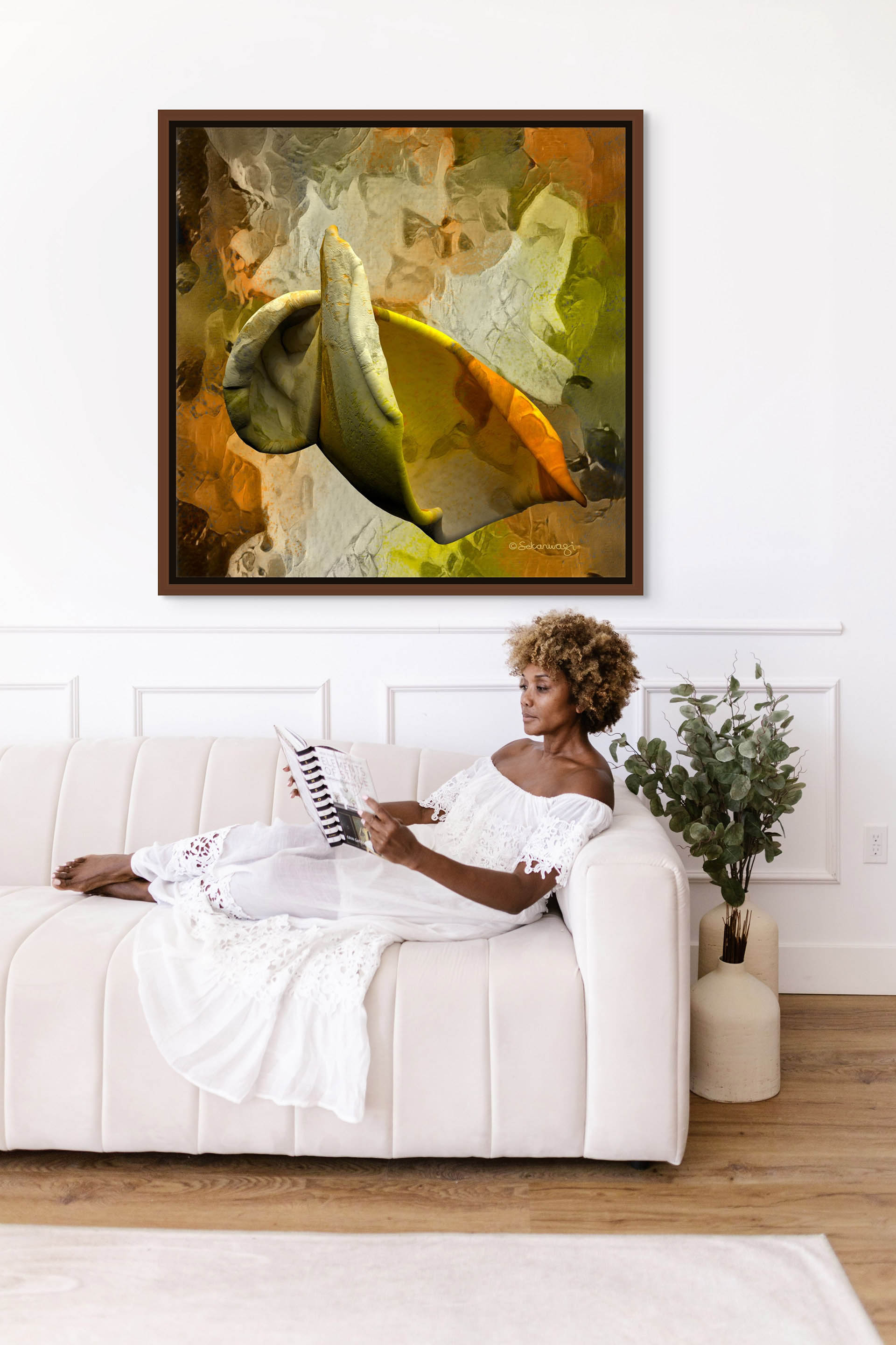 A woman in a white dress reclines on a cream sofa, reading a magazine. Behind her, the Emerging Thought - Canvas Art in warm colors adorns the wall. Beside the sofa, a green potted plant adds freshness to the minimalist room with its white walls.