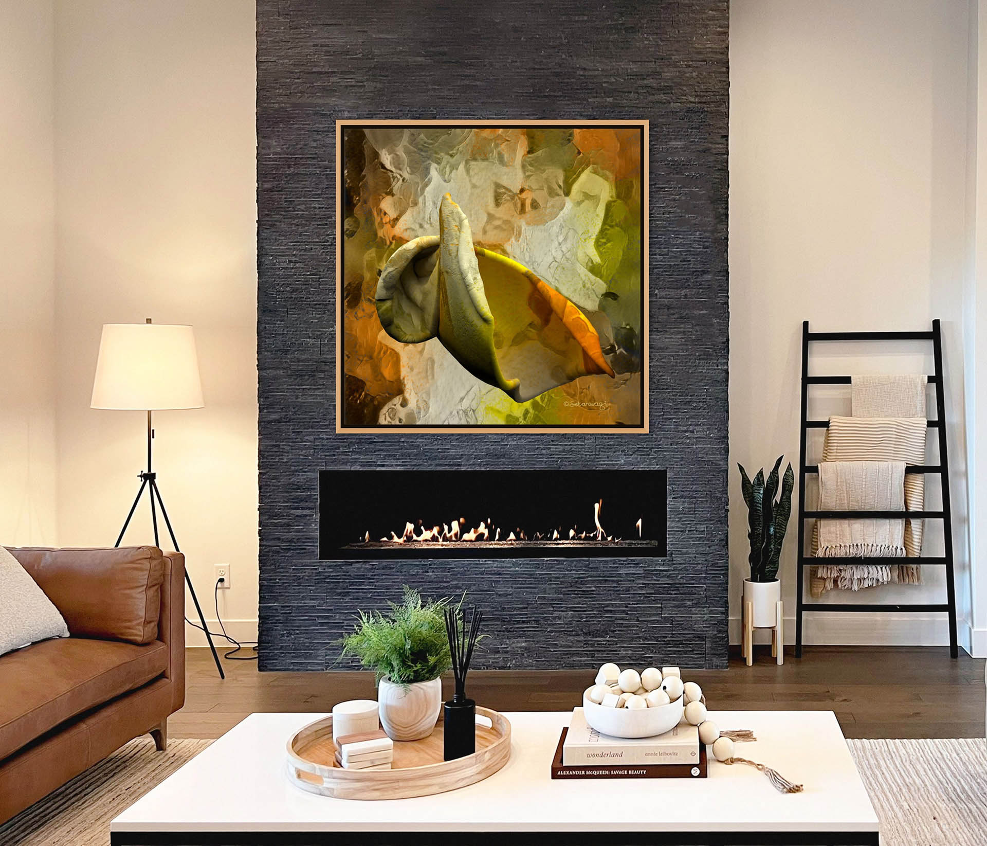 A modern living room showcases a brown leather sofa, a white coffee table with a plant and decor, and a ladder shelf holding blankets. The sleek fireplace against a dark stone wall is adorned with the Emerging Thought canvas art. A floor lamp stands to the left.