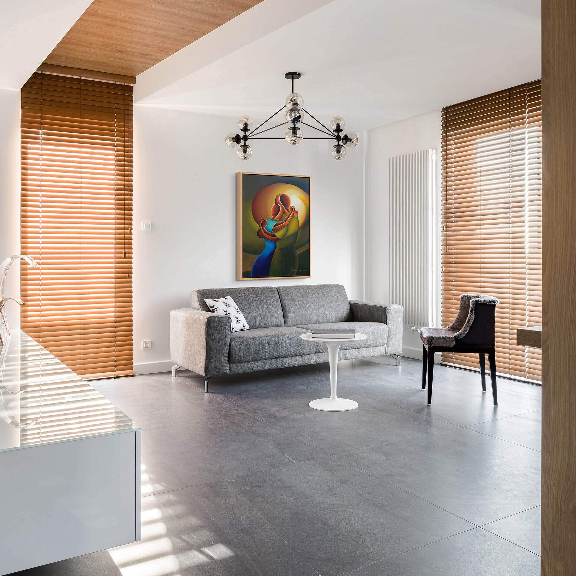 A modern living room showcases a gray sofa, Love Me Tender canvas art, and a round white table. A black accent chair is nearby. Wooden blinds cover large windows, while a unique chandelier above enhances the bright, spacious atmosphere.
