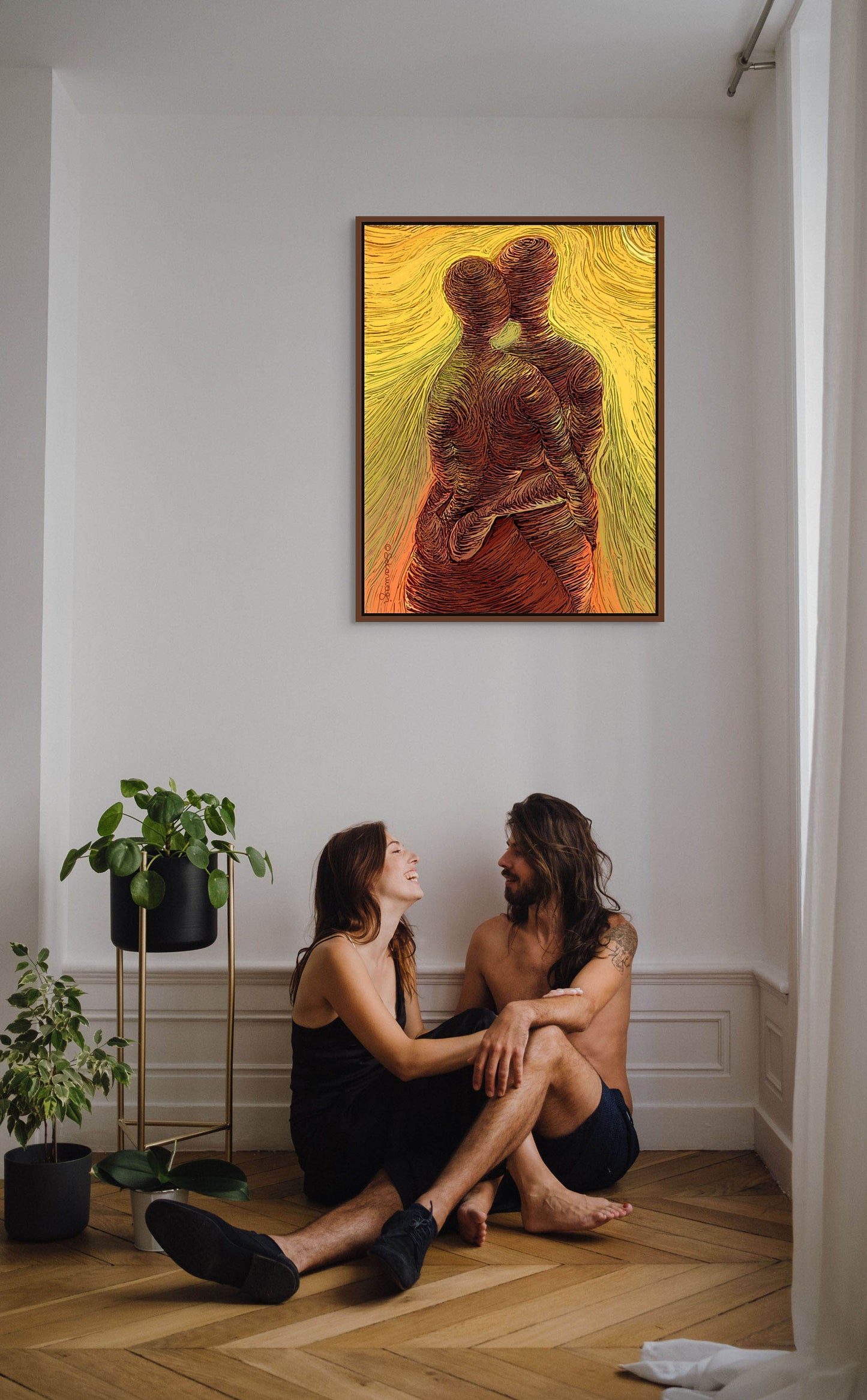 A couple sits on a wooden floor in casual wear, exchanging smiles. Next to them is a potted plant. Above, Loving Me, Loving You - Canvas Art decorates the white wall with its vibrant abstract depiction of two embracing figures, printed on premium quality cotton canvas.