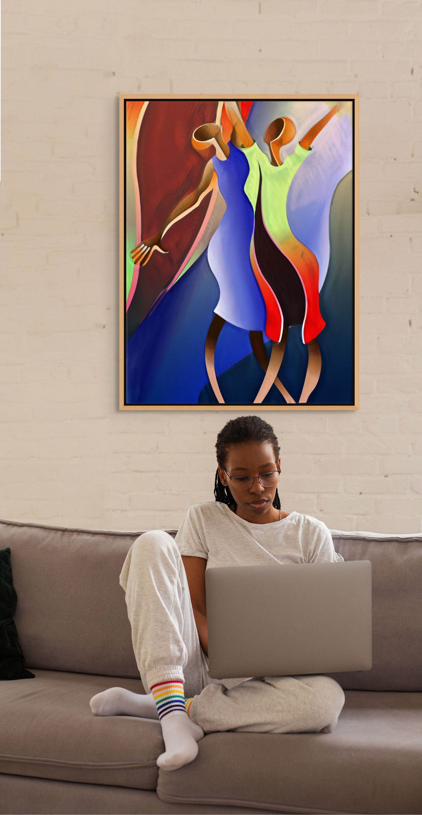 A person sits on a grey couch, working on a laptop. They wear glasses, a grey outfit, and rainbow-striped socks. Behind them is Reaching For The Sky, an exquisite abstract canvas art print featuring two dancing figures. Its ready to hang with free shipping.