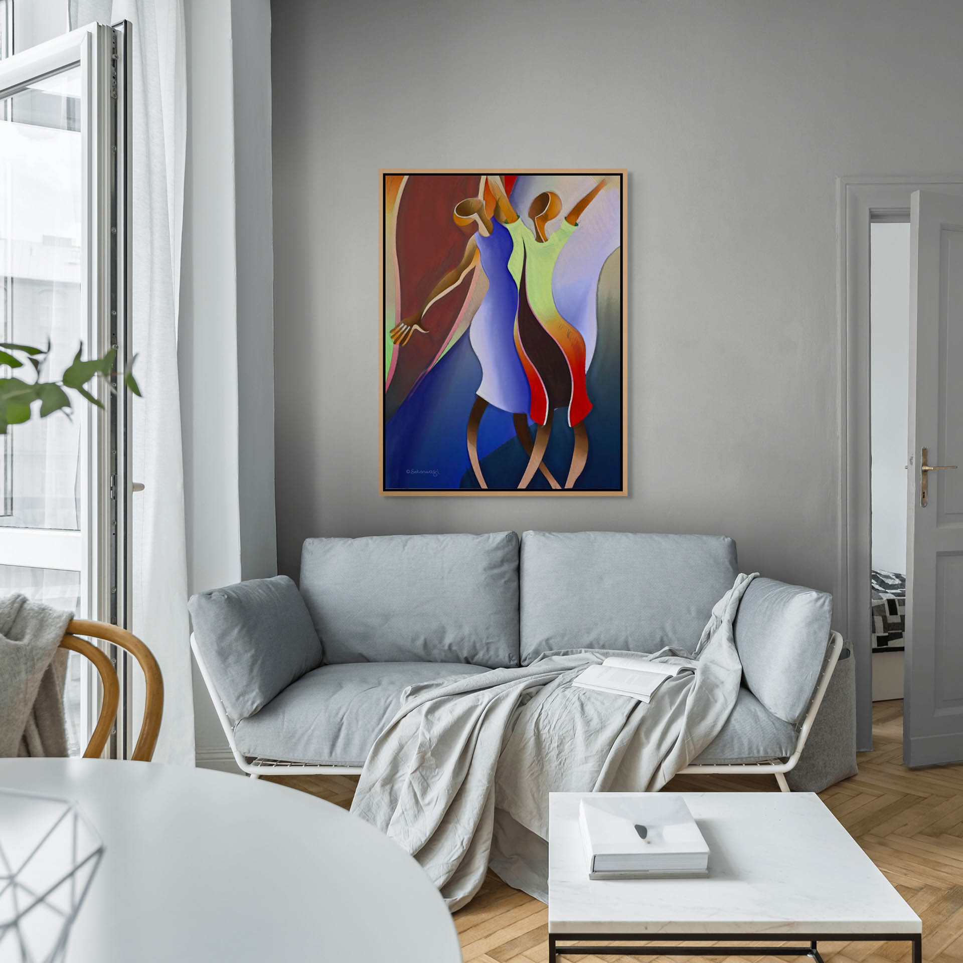 In a modern living room, a light gray sofa, white coffee table, and wooden chair set the scene. Sunlight filters through white-curtained windows, enhancing the inviting atmosphere. On the wall hangs Reaching For The Sky - Canvas Art, an abstract piece showcasing two stylized figures in vibrant colors.