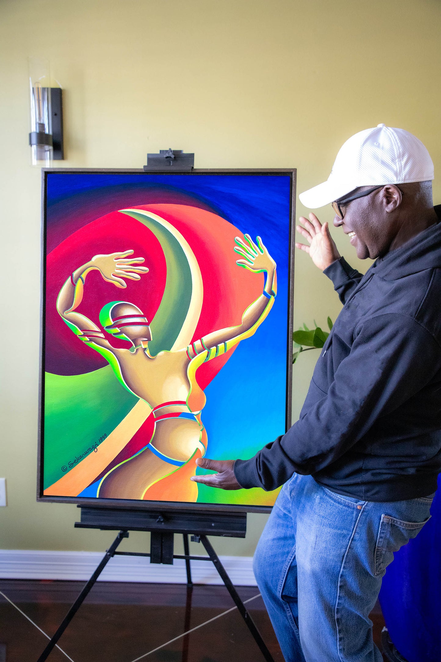 A person in a white cap and glasses stands beside The Awakening - Canvas Art on an easel, gesturing toward it. This artwork, reminiscent of Abstract African Art, features an abstract figure with raised arms amidst swirling colors.