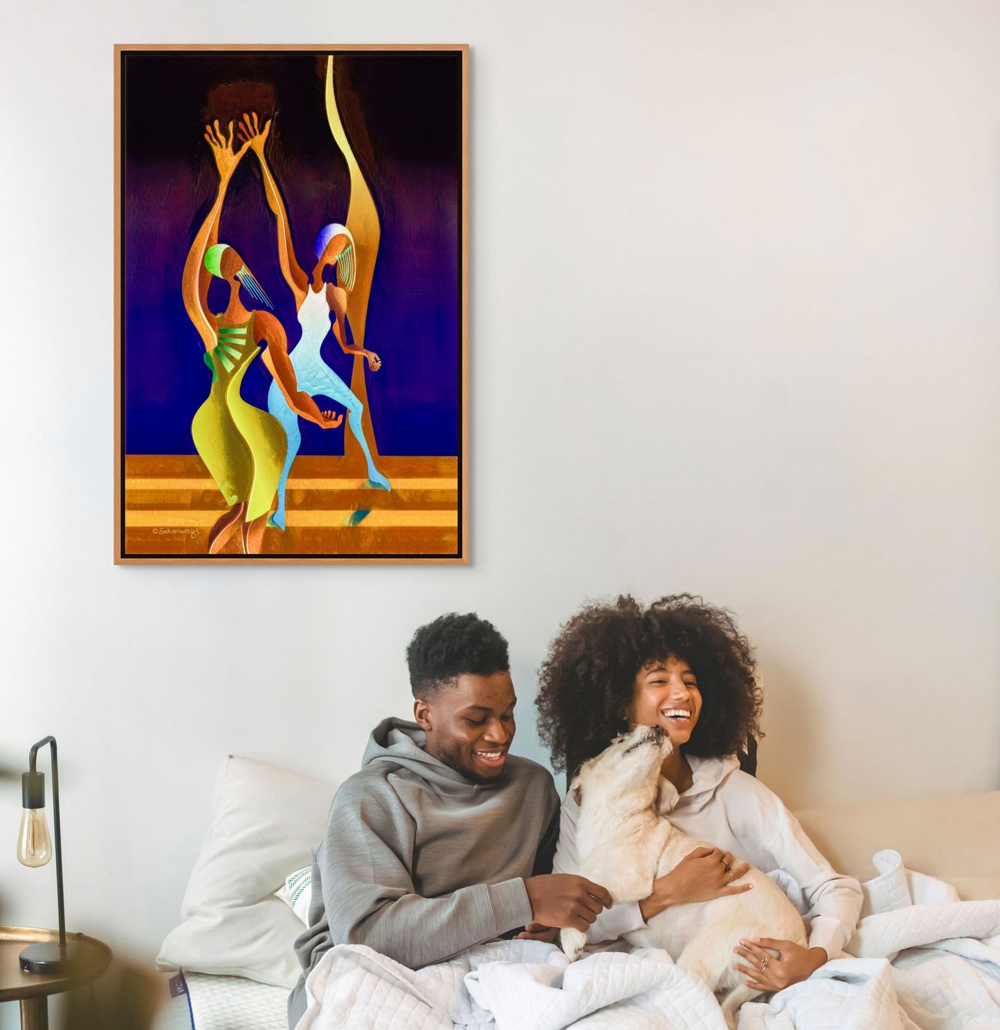 A couple joyfully sits on a bed with their dog, under the vibrant Victory Dance - Canvas Art featuring abstract dancing figures. To the left, a lamp is placed on a bedside table.