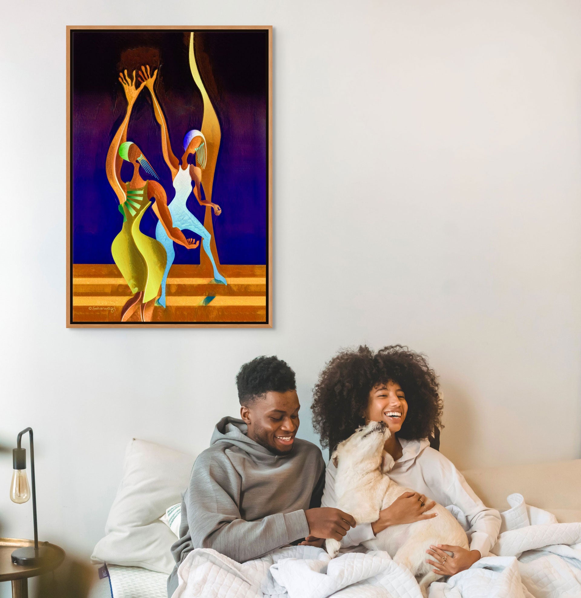 A couple joyfully sits on a bed with their dog, under the vibrant Victory Dance - Canvas Art featuring abstract dancing figures. To the left, a lamp is placed on a bedside table.
