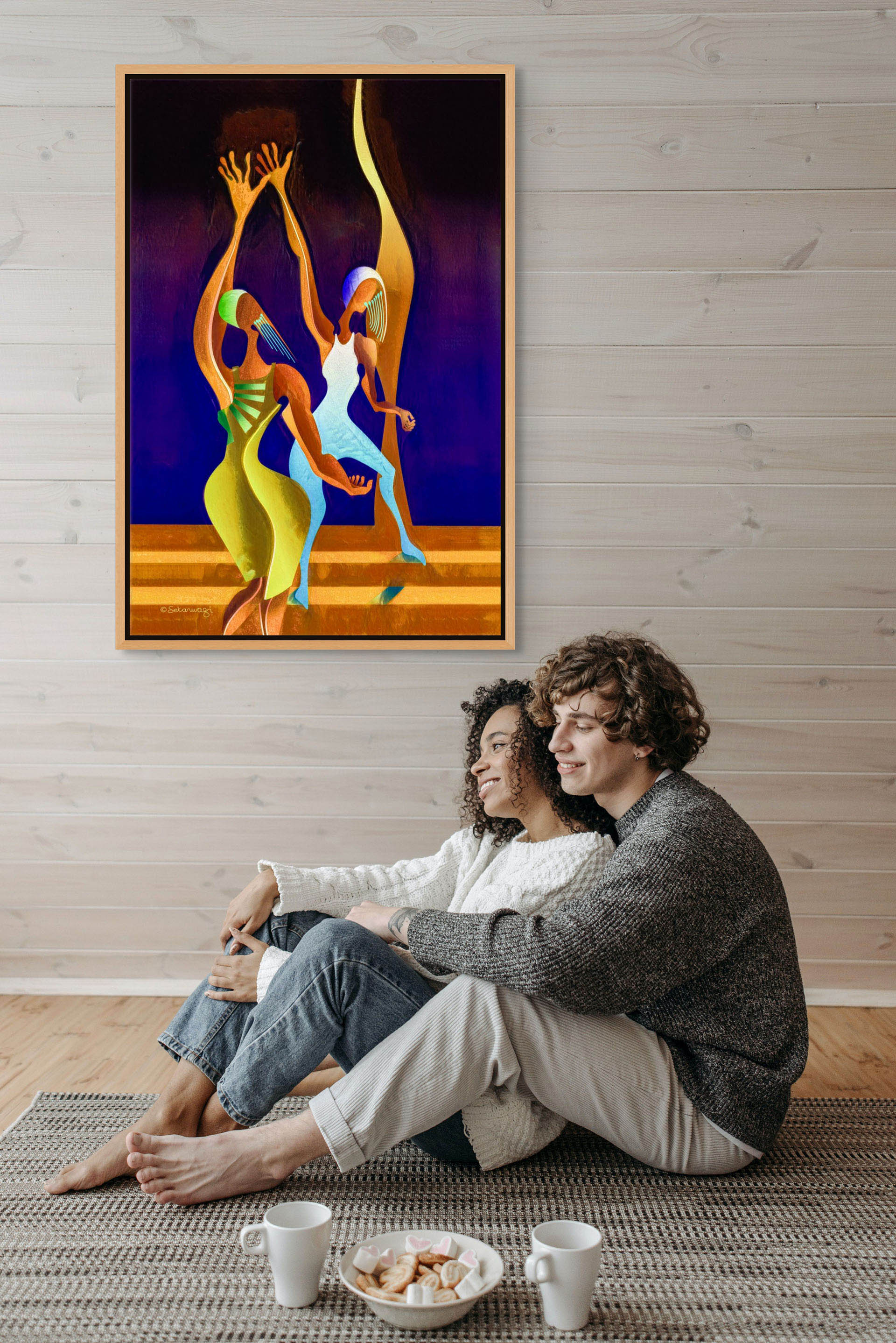 In a snug, light-wood room, a couple admires the Victory Dance canvas art on the wall, featuring vibrant, fluid human figures against a deep blue backdrop. They sit on a rug with two mugs and a plate of cookies.